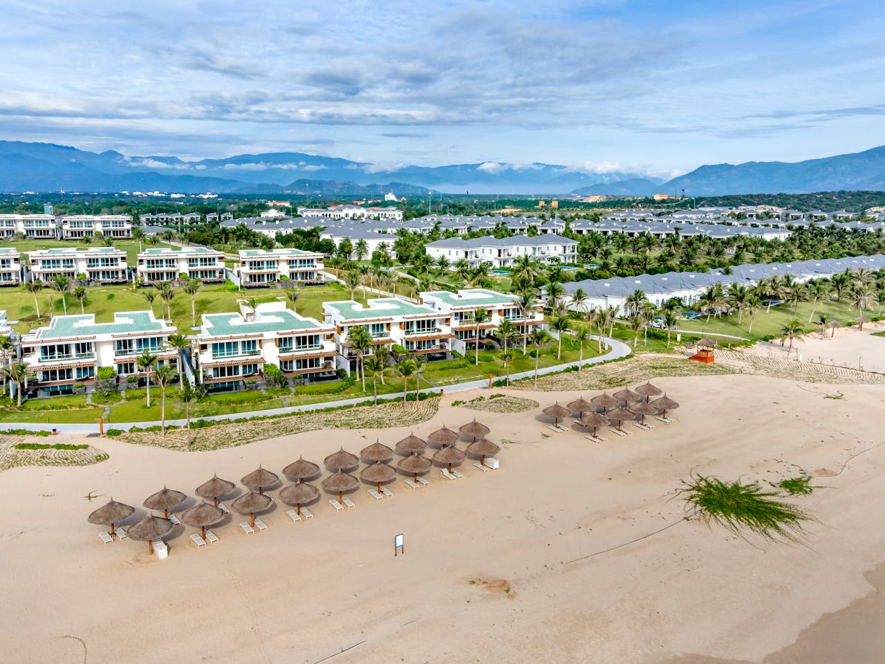 Aerial shot of beachfront with umbrella huts, luxury resort, and mountains in the background.